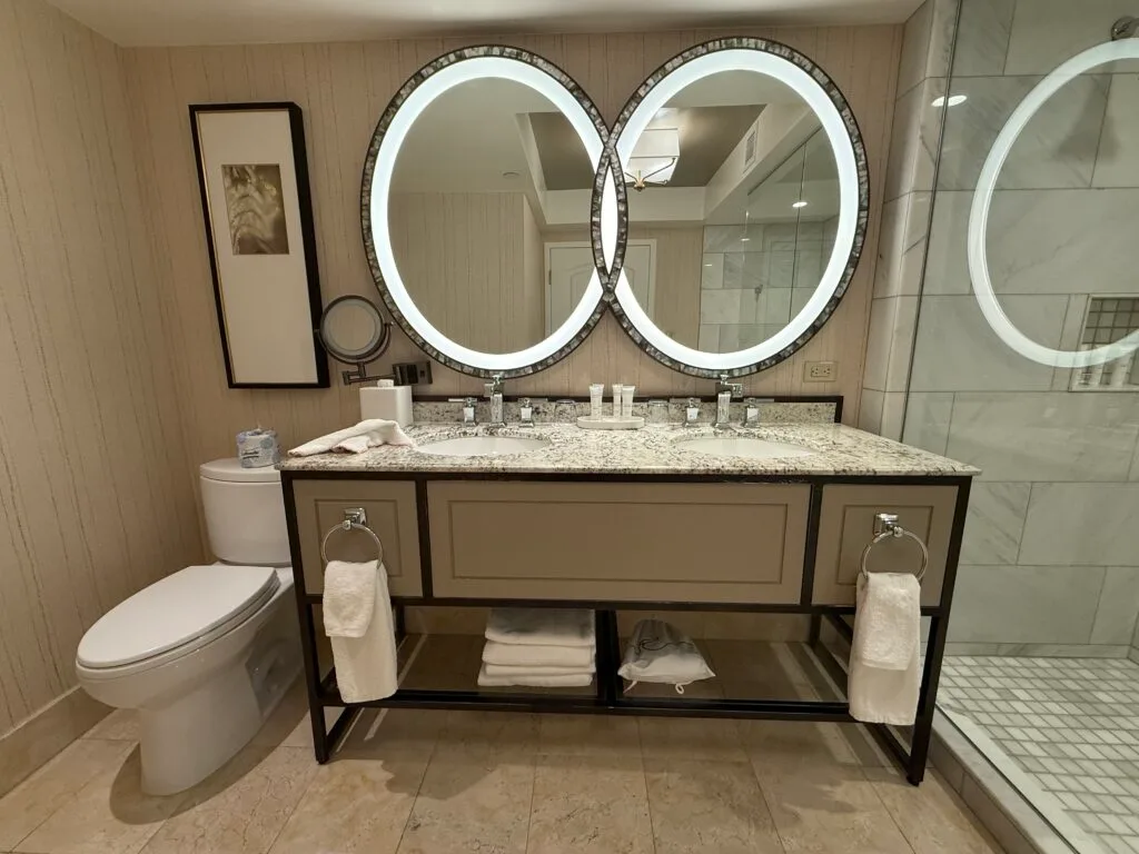 A dual sink vanity topped with an  illuminated mirrr wihch appears to combine two circular mirrors into one. 