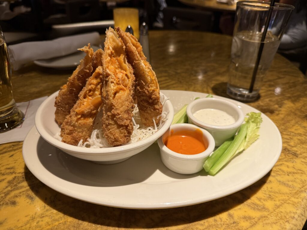 Several buffalo chicken rolls set up upright, resembling a small teepee on a plate next to tubs of ranch and buffalo sauce.