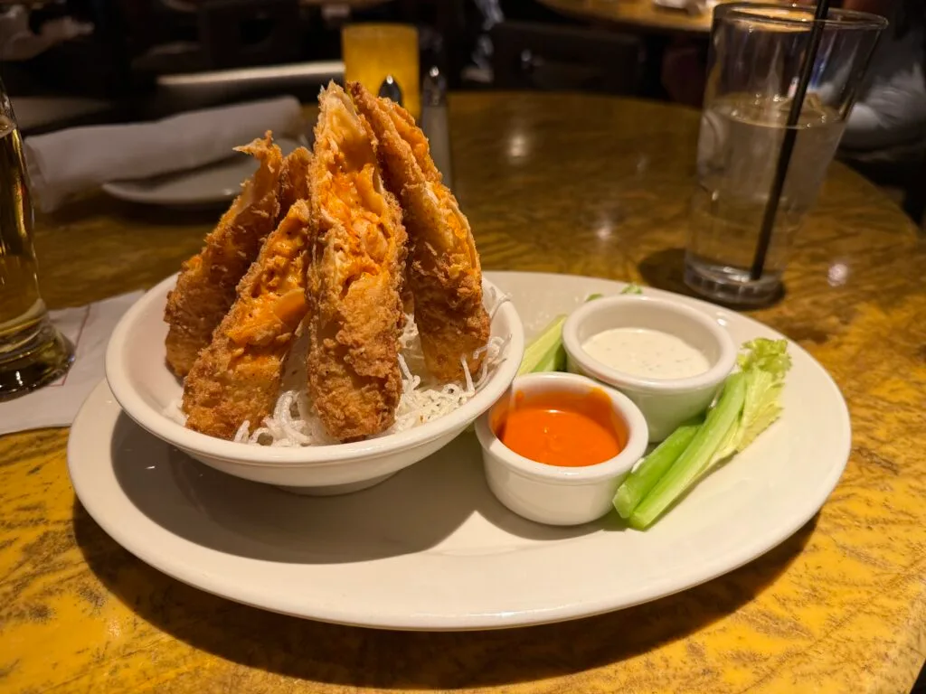 Several buffalo chicken rolls set up upright, resembling a small teepee on a plate next to tubs of ranch and buffalo sauce. 