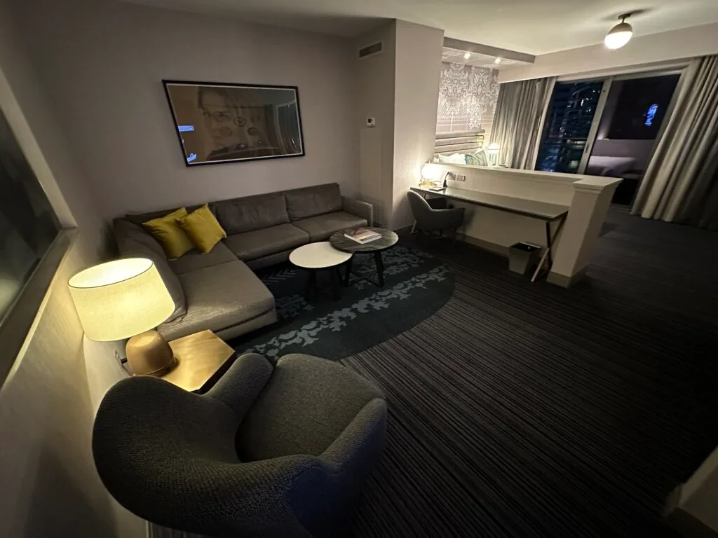 A dark living area with a chair, couch, and coffee table. The bedroom area is partially separated and visible in the background. 