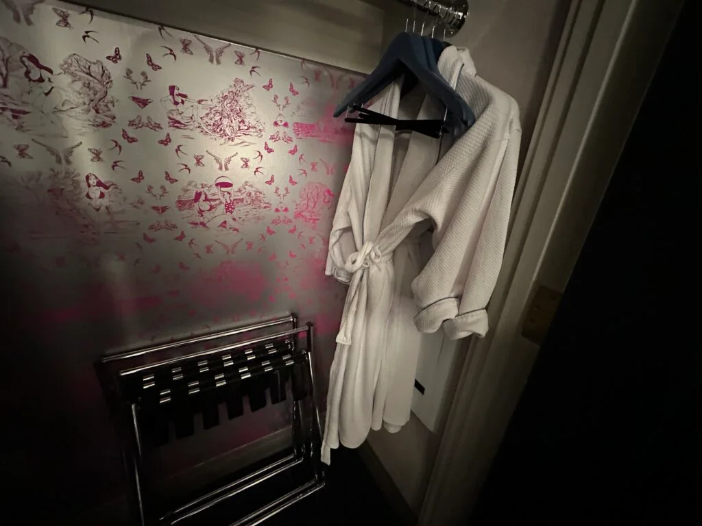 Two white robes hanging in an illuminated closet with a pink, silver metallic wallpaper on the back wall. 