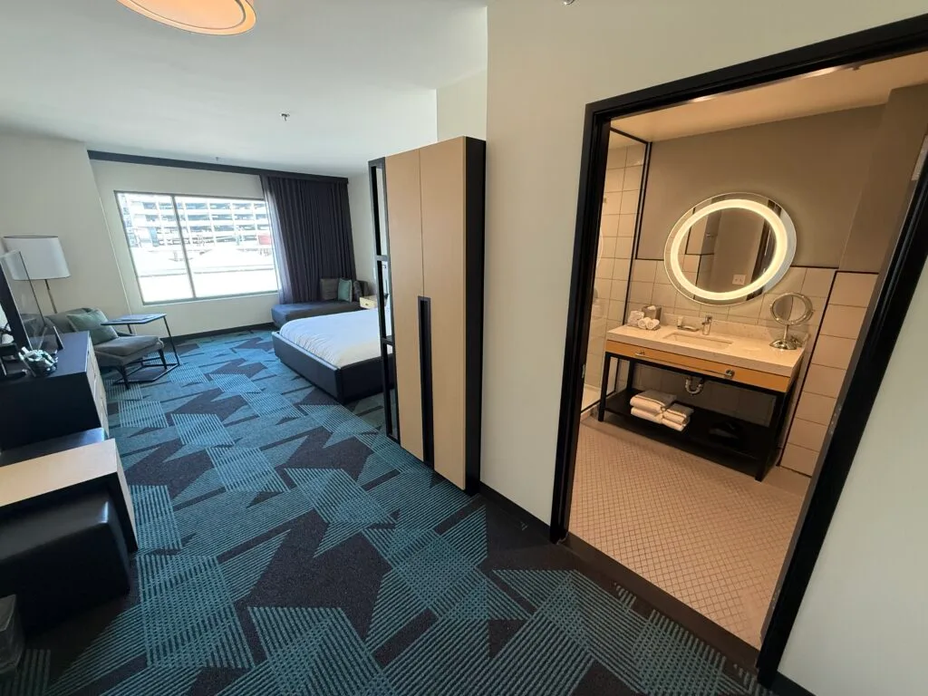 A shot from the entry way of the door leading to the bathroom, with an illuminated mirror visible, and the room in the background. 