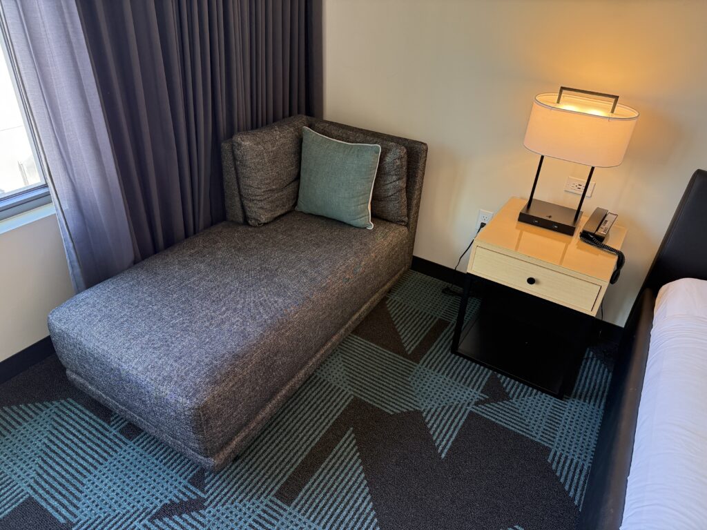 A small couch in the corner, with one decoratove pillow on it.