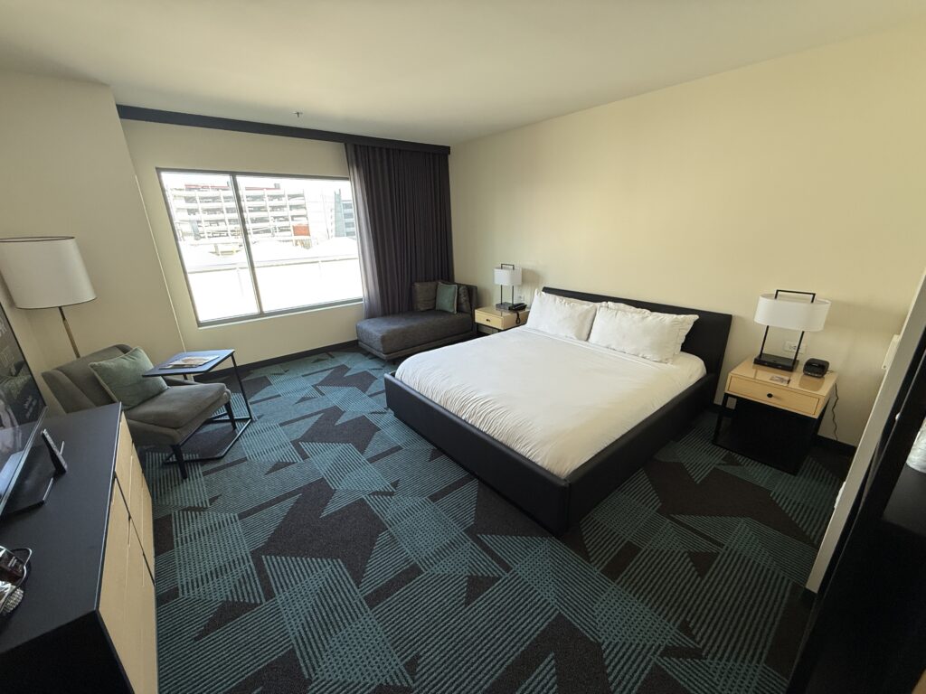 A wide angle of the room, with a king bed, teal carpet, and a window in the background.