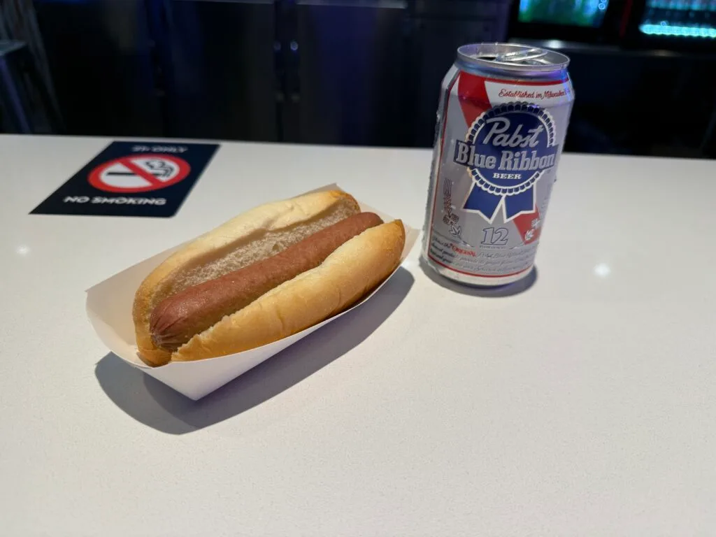 A hot dog, on a table next to a can of PBR. 