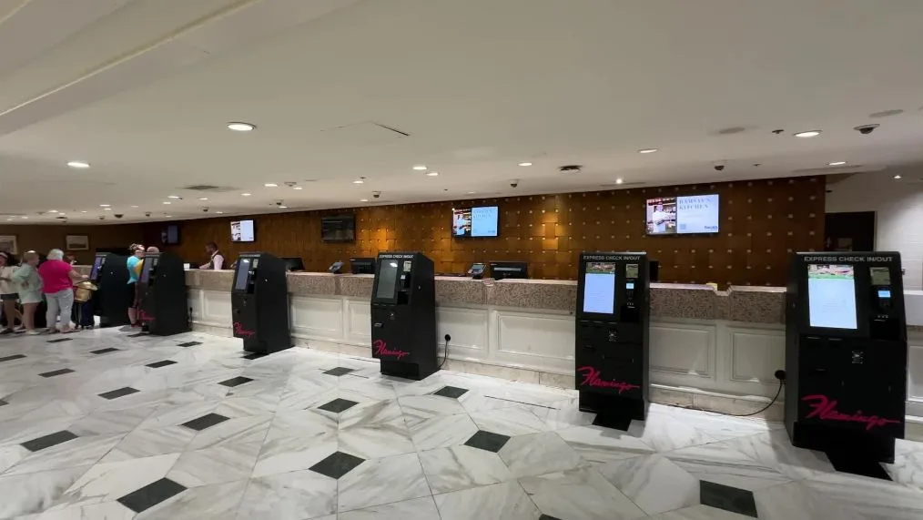 A bank of self check in kiosks stand vacant in front of the lobby desk. 
