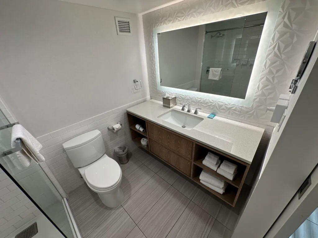 A single sink vanity with a backlit mirror above it. A toilet is crammed between the vanity and the shower . 