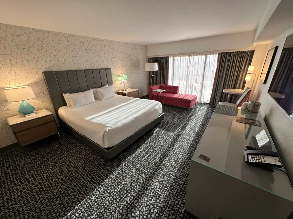 A wide angle shot of the room showing a king bed in the middle of the room, with the windows on the back wall. 