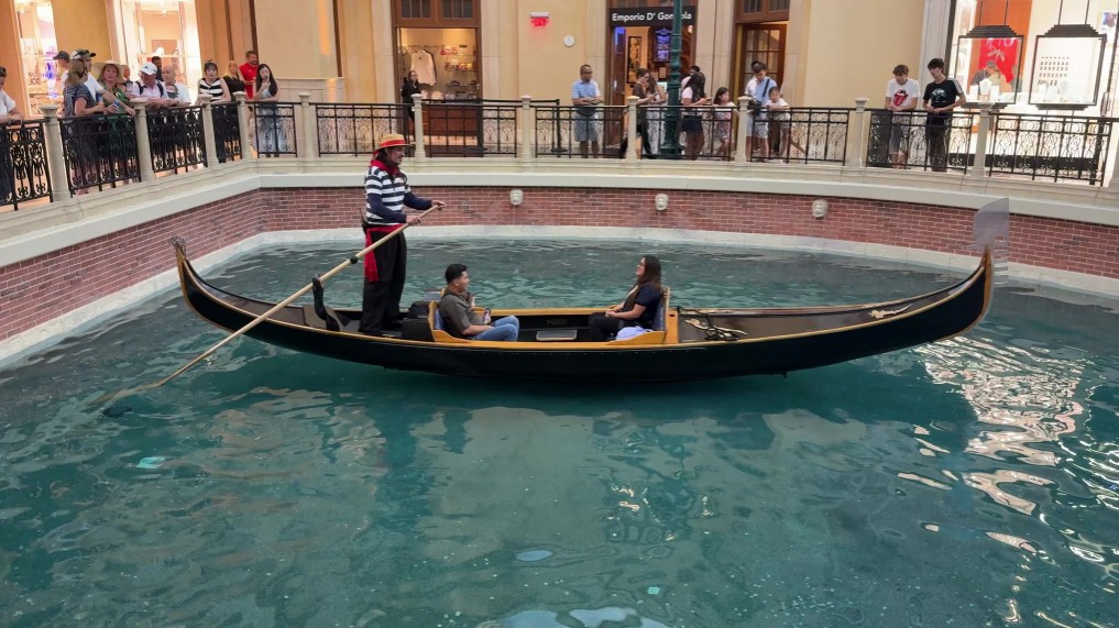 A gondola with 2 passnegers navigating a canal. The Gondolier is singing a song to them.