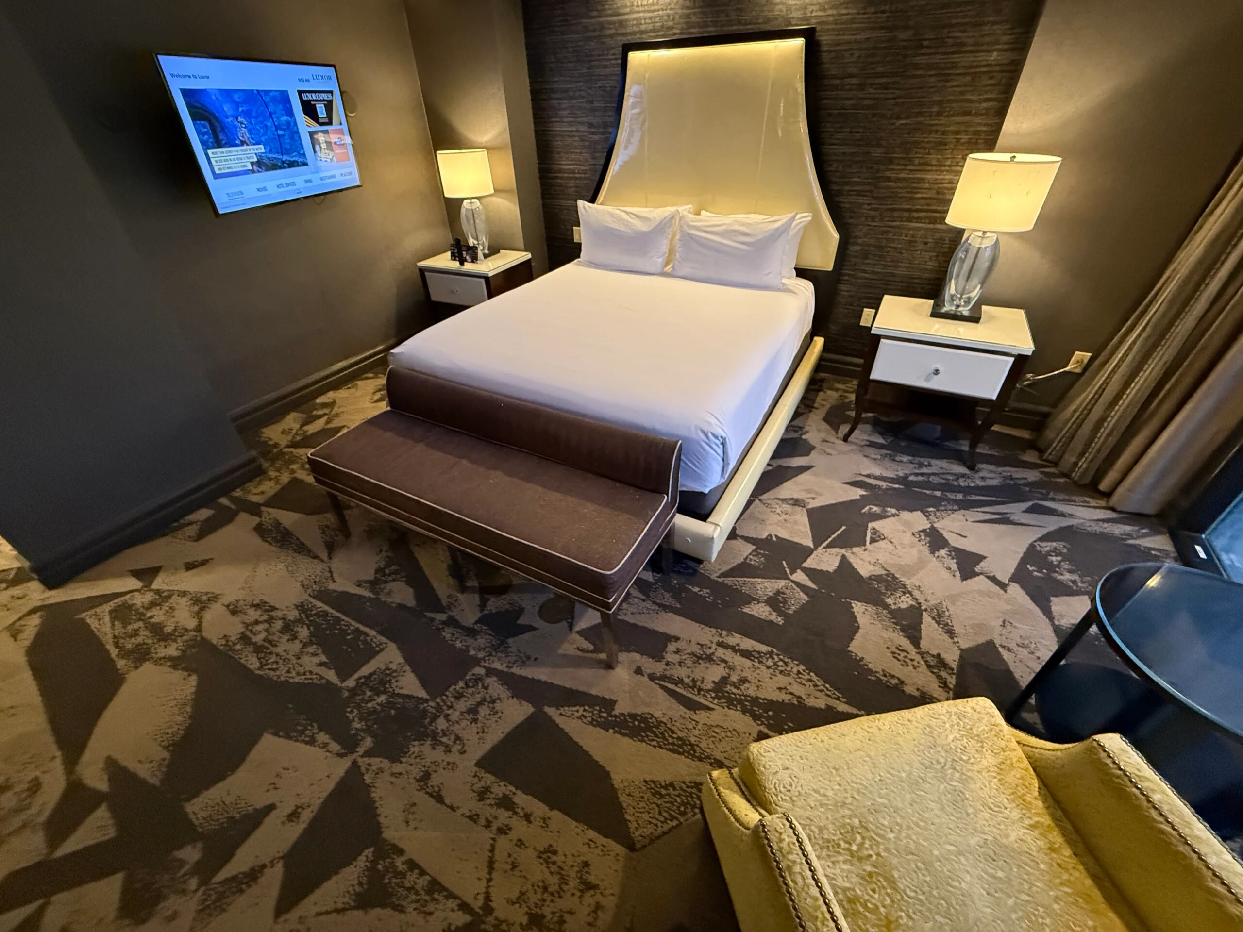 A king bed, illuminated from above, with a TV hanging on the wall to the left of it.