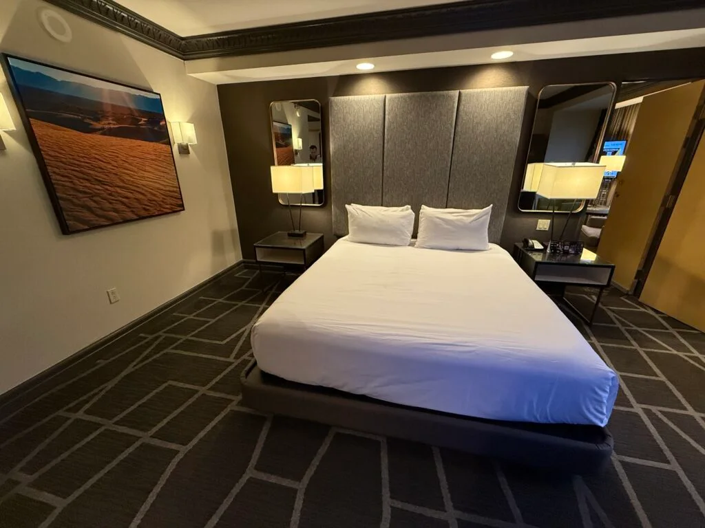 The king bed, with two lights shining down from the ceiling on it. To the left, a picture of a sand dune hangs on the wall.