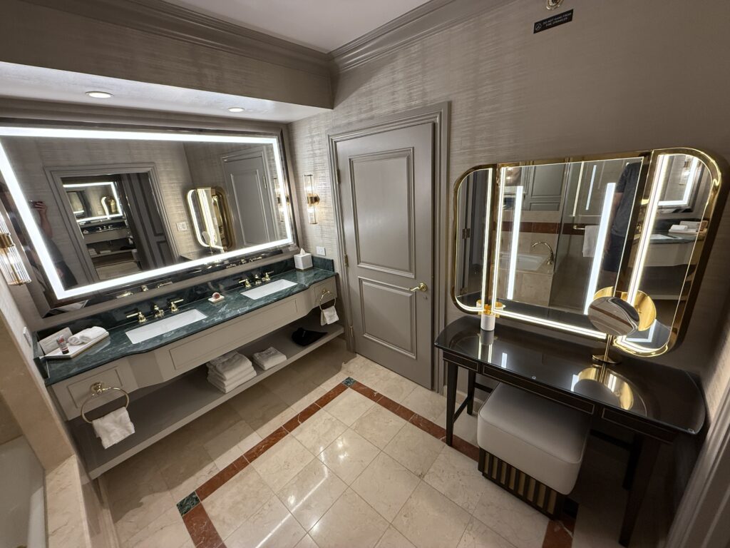 A wide angle picture of the dual sink vanity topped by an illuminated mirror, the door to the WC, and a powder station.