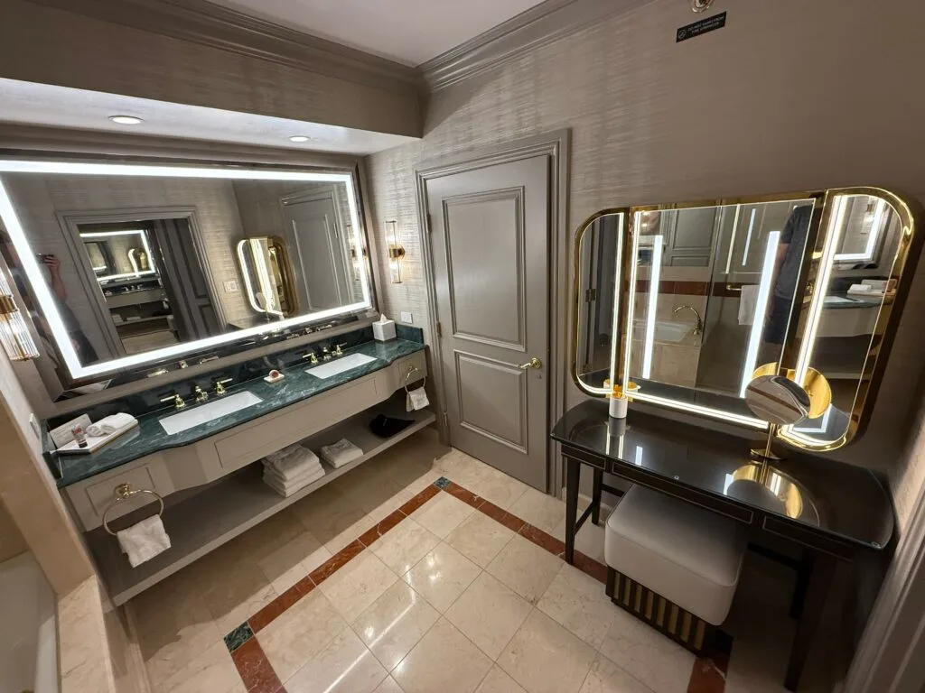 A wide angle picture of the dual sink vanity topped by an illuminated mirror, the door to the WC, and a powder station. 