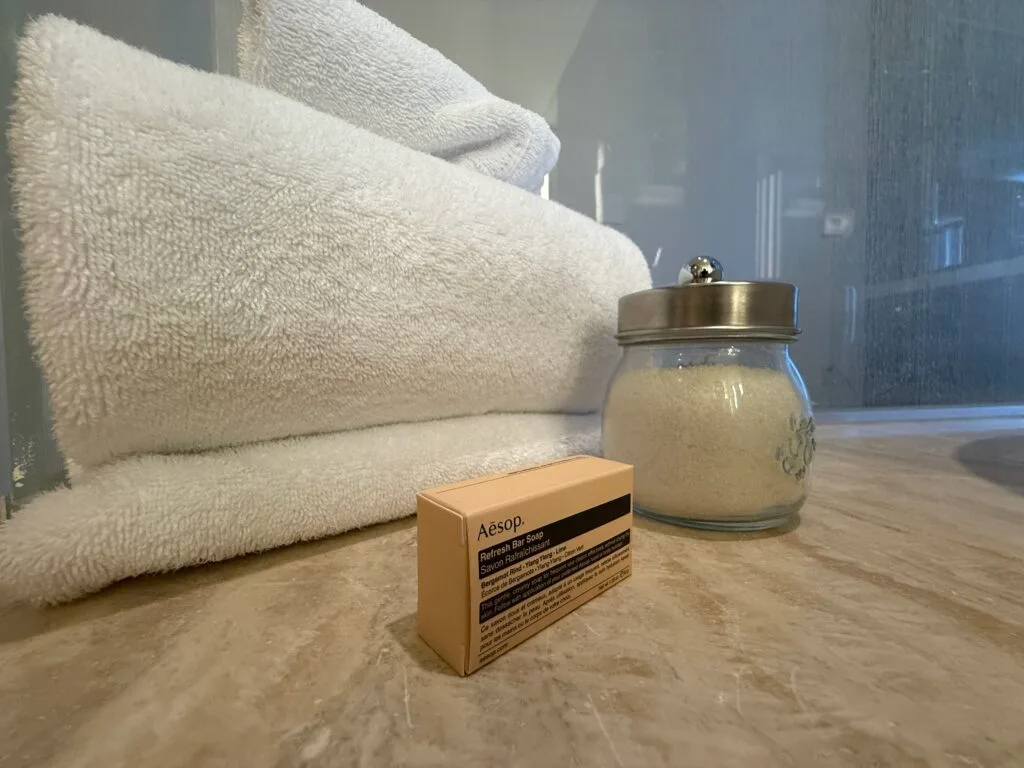 A glass jar full of salt next to a box of soap, both positioned in front of a stack of folded towels. 