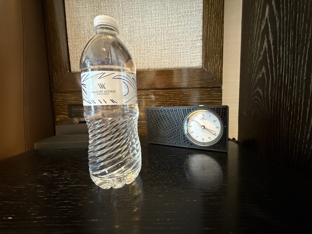 A 12oz bottle of water on an end table next to an analog clock radio.
