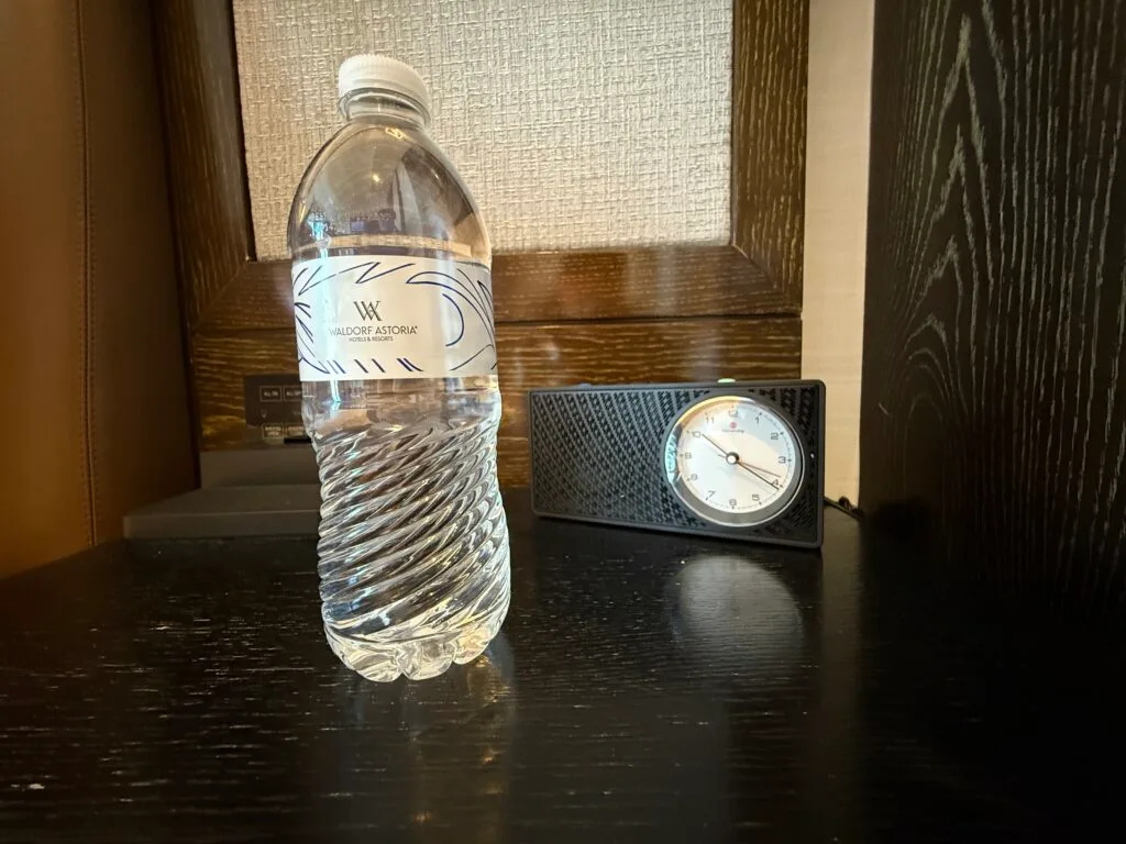 A 12oz bottle of water on an end table next to an analog clock radio. 