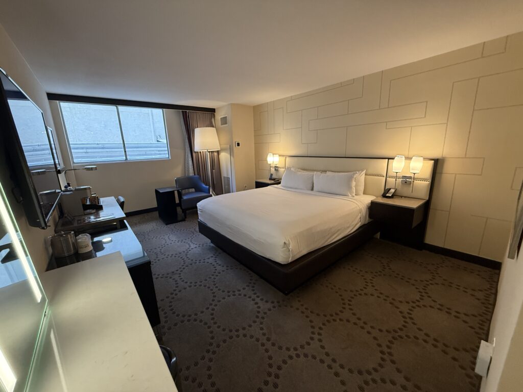A wide angle shot of the room, with a king bed up against a wall and a small window in the background.