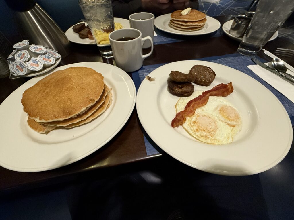 Two plates on a table, one with 3 pancakes, and the other with a slice of bacon, 2 sausage patties and two eggs.