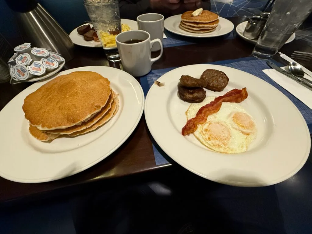 Two plates on a table, one with 3 pancakes, and the other with a slice of bacon, 2 sausage patties and two eggs. 