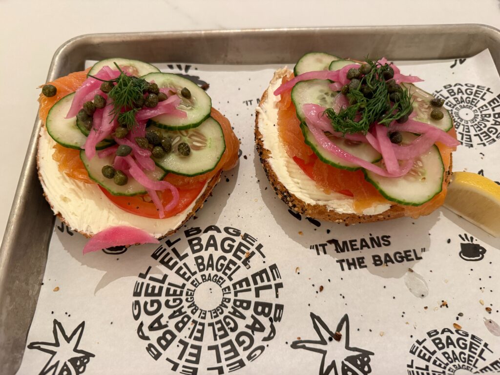 Two halves of a bagel, laying on a tray stacked with salmon, and veggies.