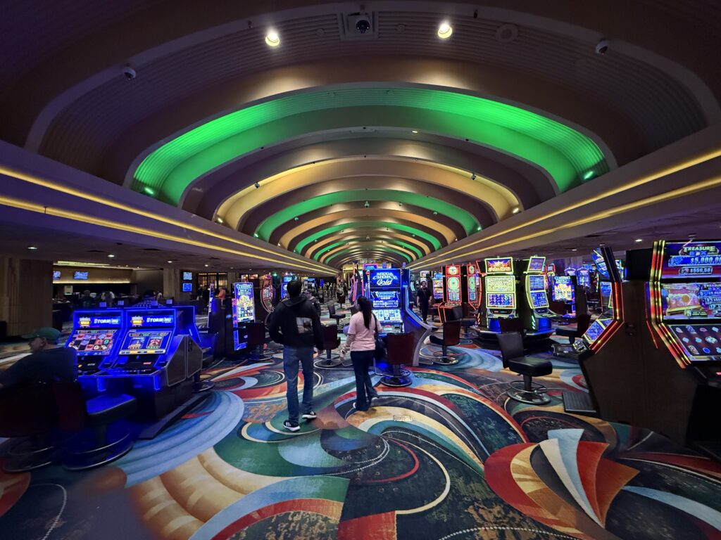 A portion of the casino floor filled with slot machines with green and yellow lighting on the ceiling. 