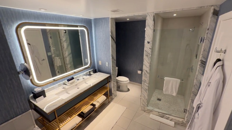A dual faucet vanity, topped by an illuminated mirror, next to the water closet and toilet and a single stall shower.