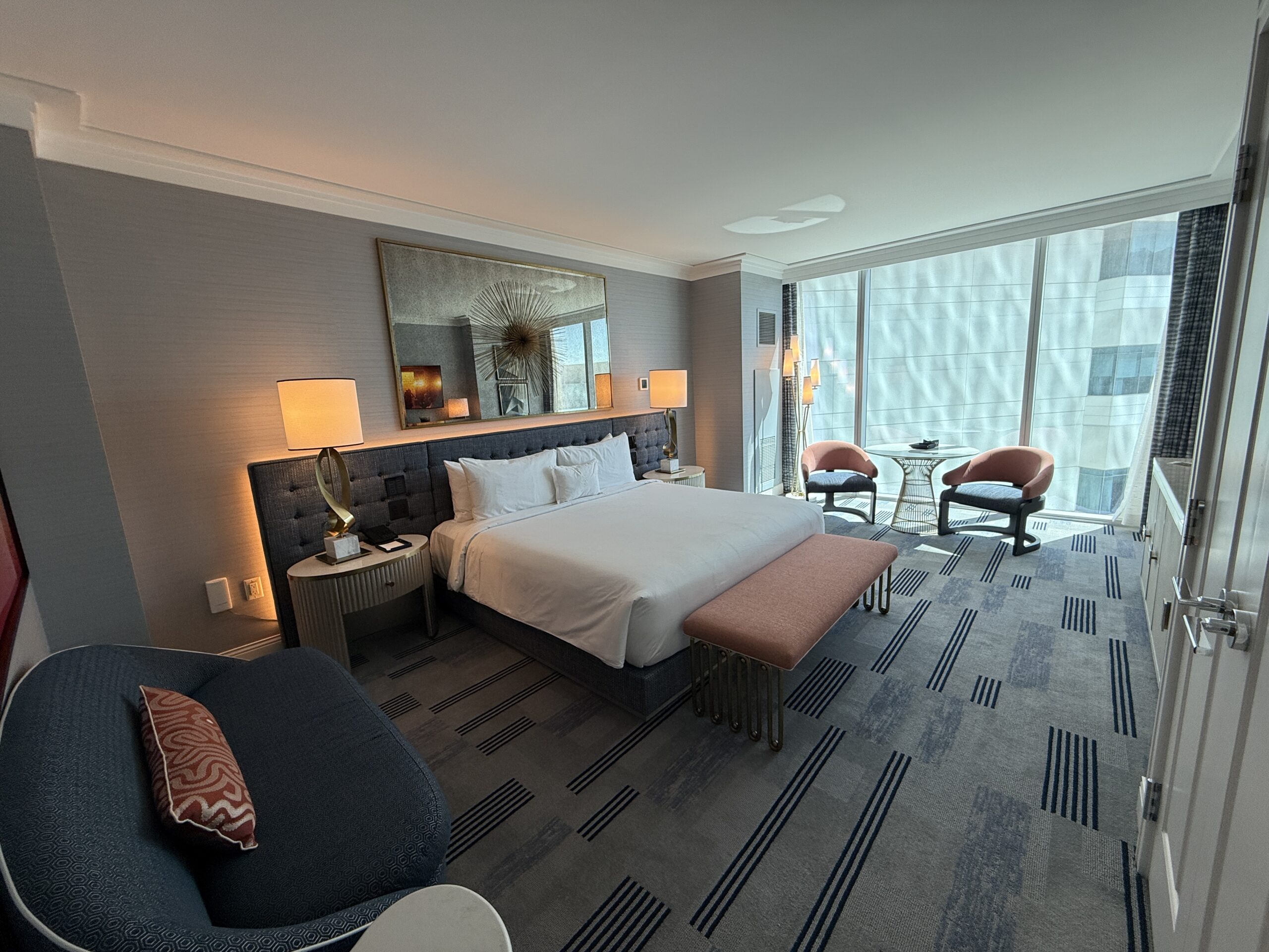 A wide angle of the room, with the king bed featured with a bench at the end of it. Window in the background.