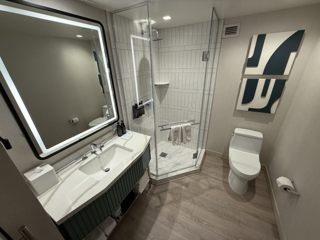 A small bathroom with an illuminated mirror atop a single sink vanity, a glass enclosed shower in the corner, and a toilet to the right. 
