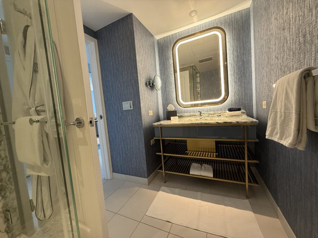 Modern bathroom with marble-topped vanity, backlit rectangular mirror, blue textured walls, and glass shower to the left.