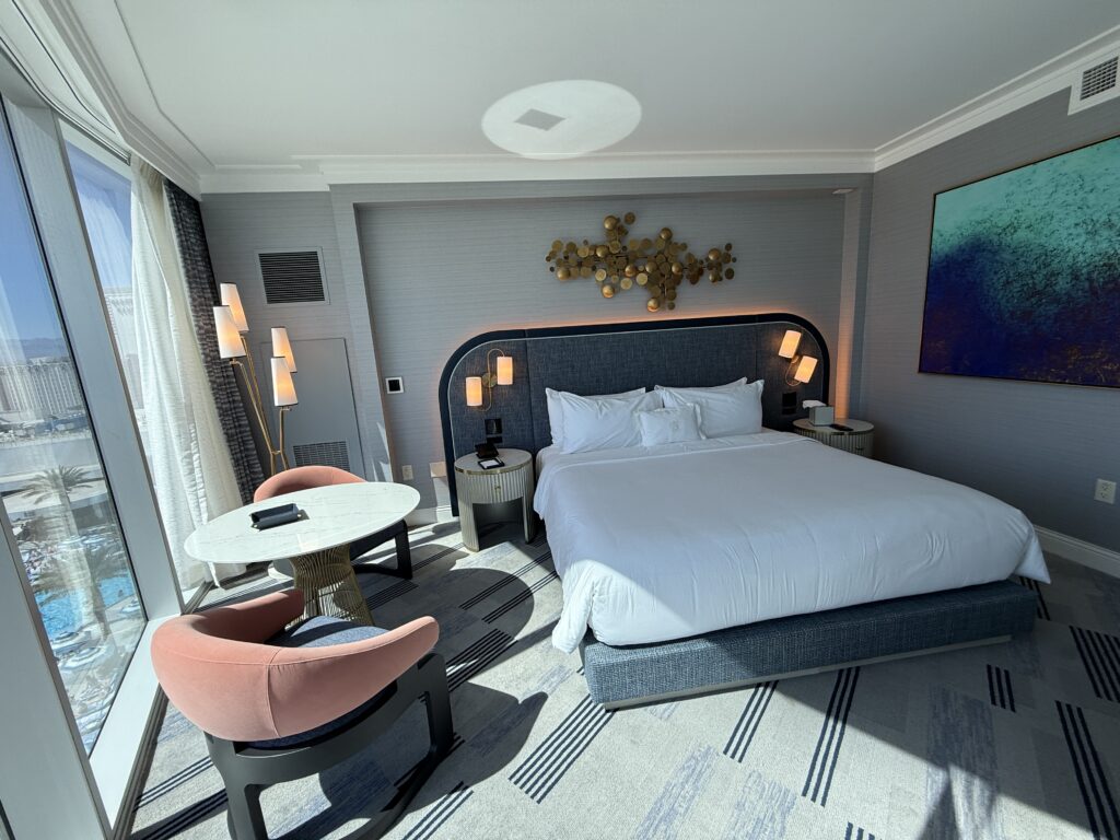 A king bed with an illuminated headboard sits next to a small table with two pink chairs. 