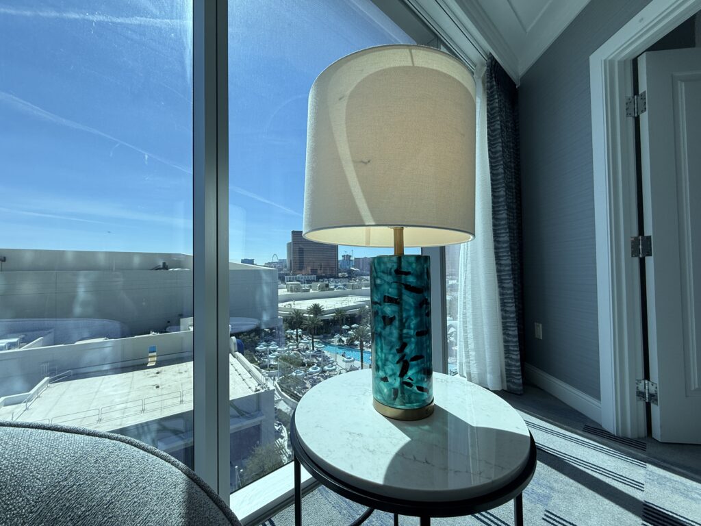 Lamp with a turquoise marbled base on a round marble-top table by a large window overlooking a hotel pool and Strip skyline outside.
