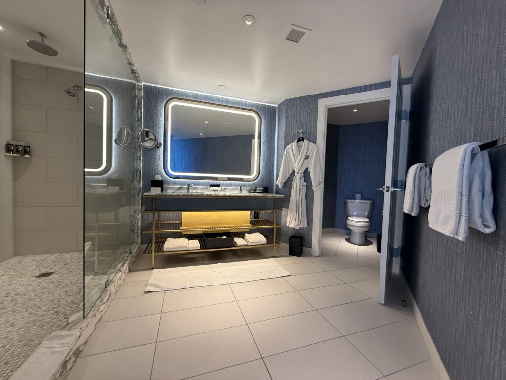 An illuminated mirror sits atop a dual sink vanity. A shower is to the left and a separate water closet with a toilet to the right. 