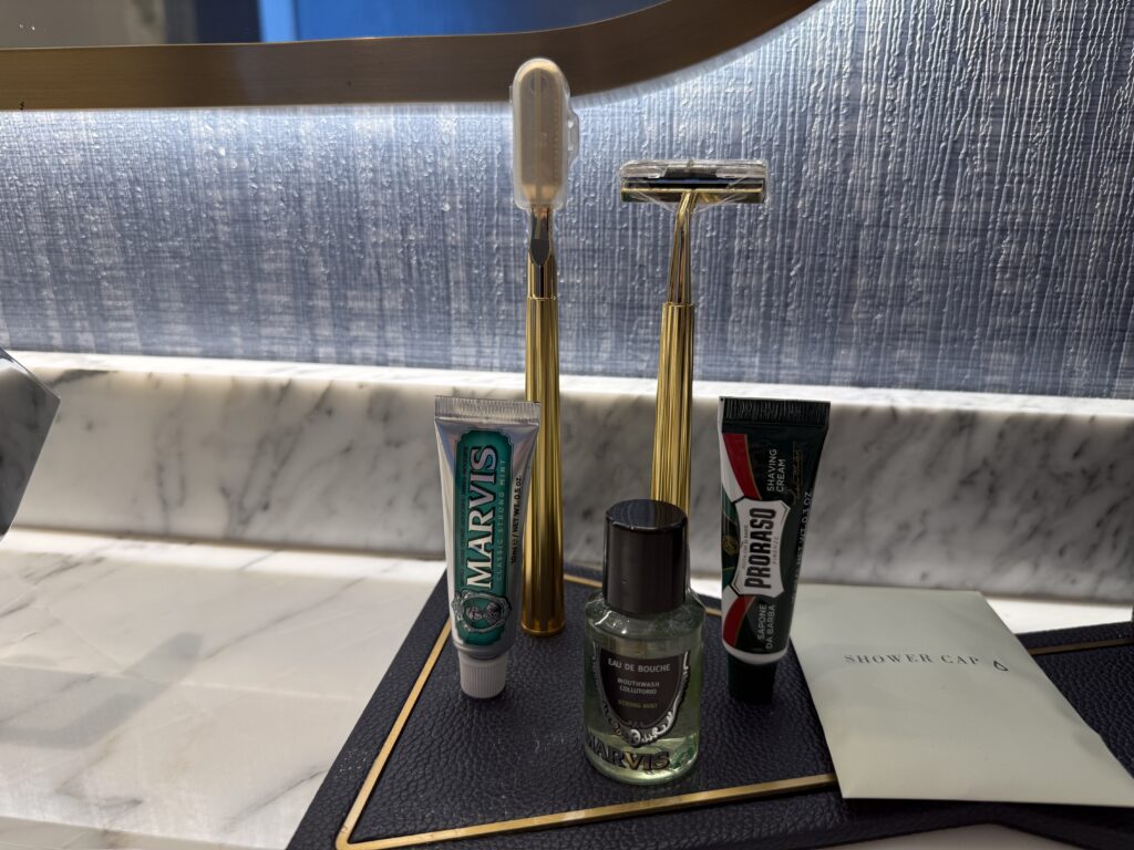 Bathroom vanity with a gold-toned razor stand holding a white razor, plus Marvis toothpaste tube, Proraso shaving cream tube, a small bottle of mouthwash, and a shower cap packet on a marble counter