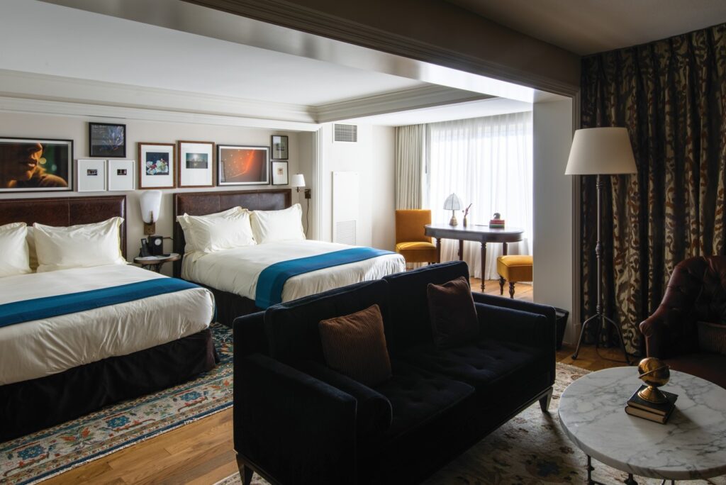 A room with two queen beds against a wall with trendy pictures hanging above the headboards. A couch is in the foreground in front of a table. 