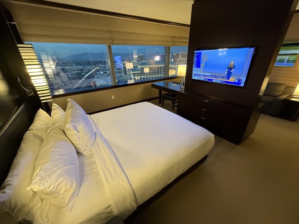 Image of the king bed positioned facing the second of the room's TVs with the windows in the background. 