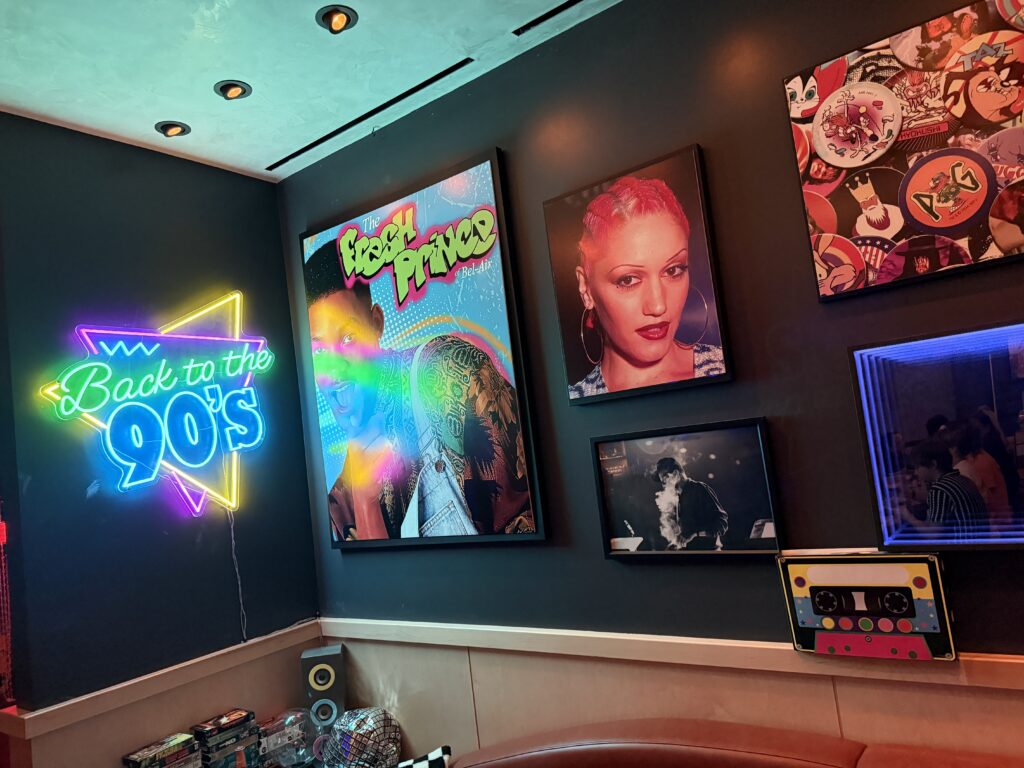 Neon sign reading 'Back to the 90s' beside a colorful The Fresh Prince of Bel‑Air poster on a dark wall.