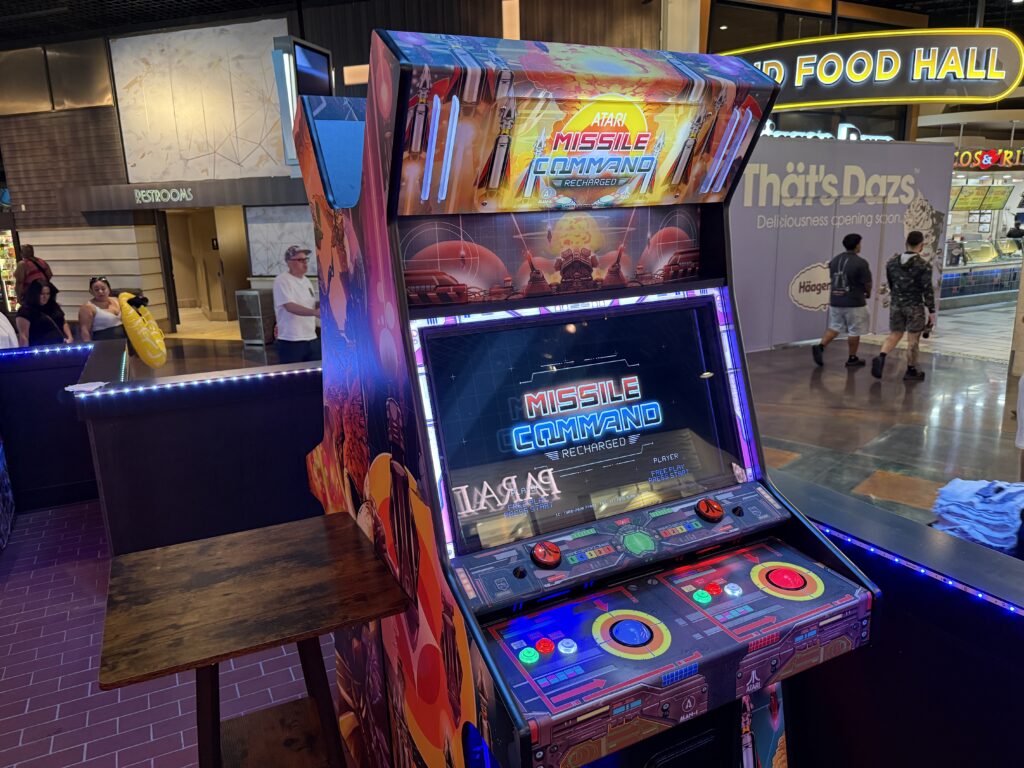 Arcade Missile Command machine in a mall, colorful cabinet and glowing screens amid other storefronts and people nearby.