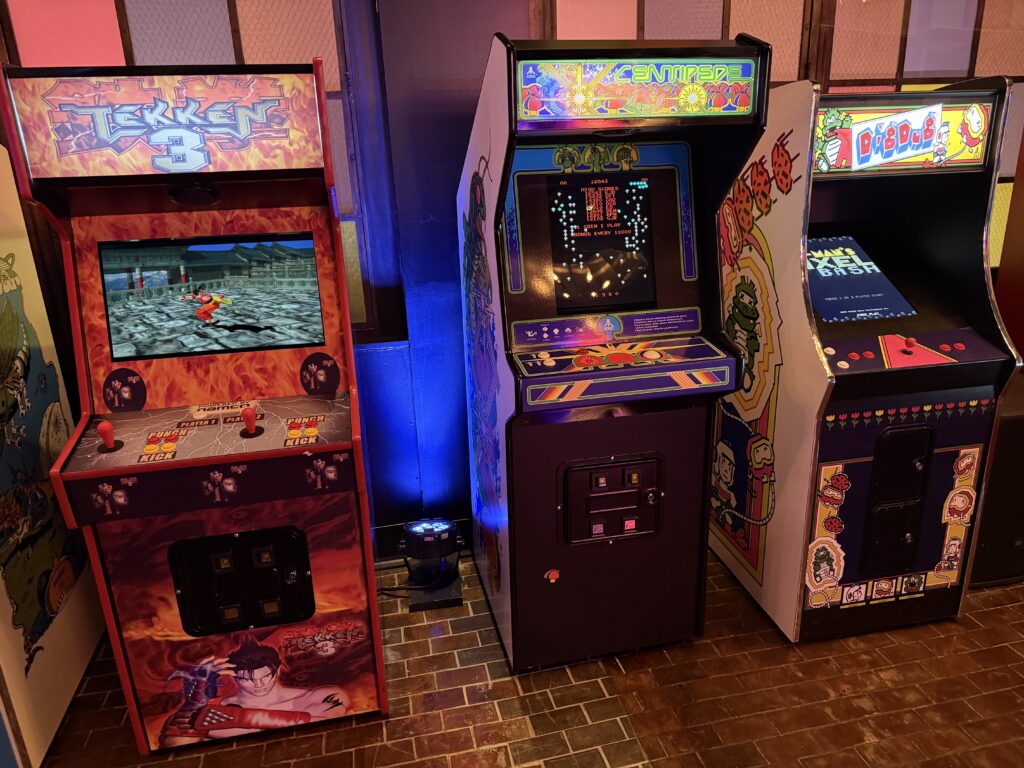 Row of colorful vintage arcade cabinets in an arcade, with screens showing gameplay and retro artwork around each cabinet.