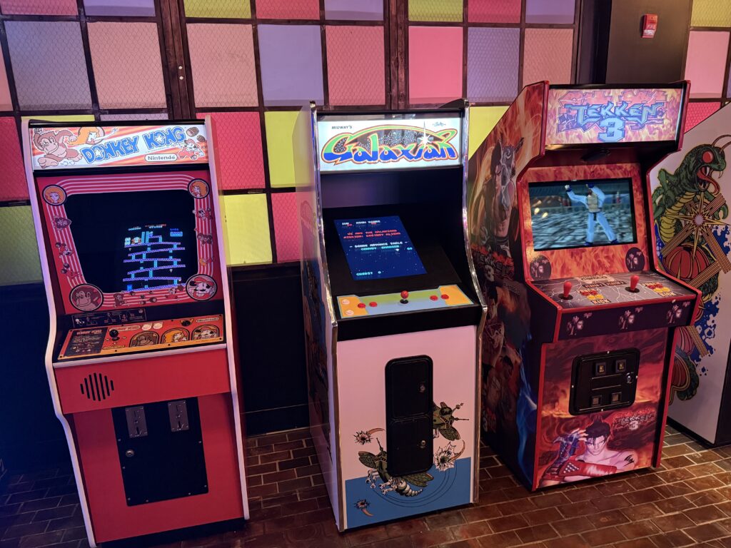 Row of vintage arcade cabinets with colorful panels: Donkey Kong on the left, Centipede in the middle, and Tekken 3 on the right, in a neon-lit arcade.
