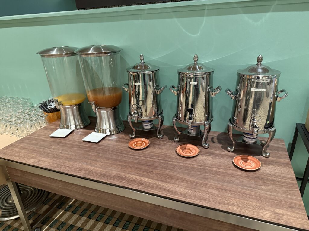 Self-serve drink station with a juice dispenser, three polished beverage urns, and a row of glasses on a wooden counter.