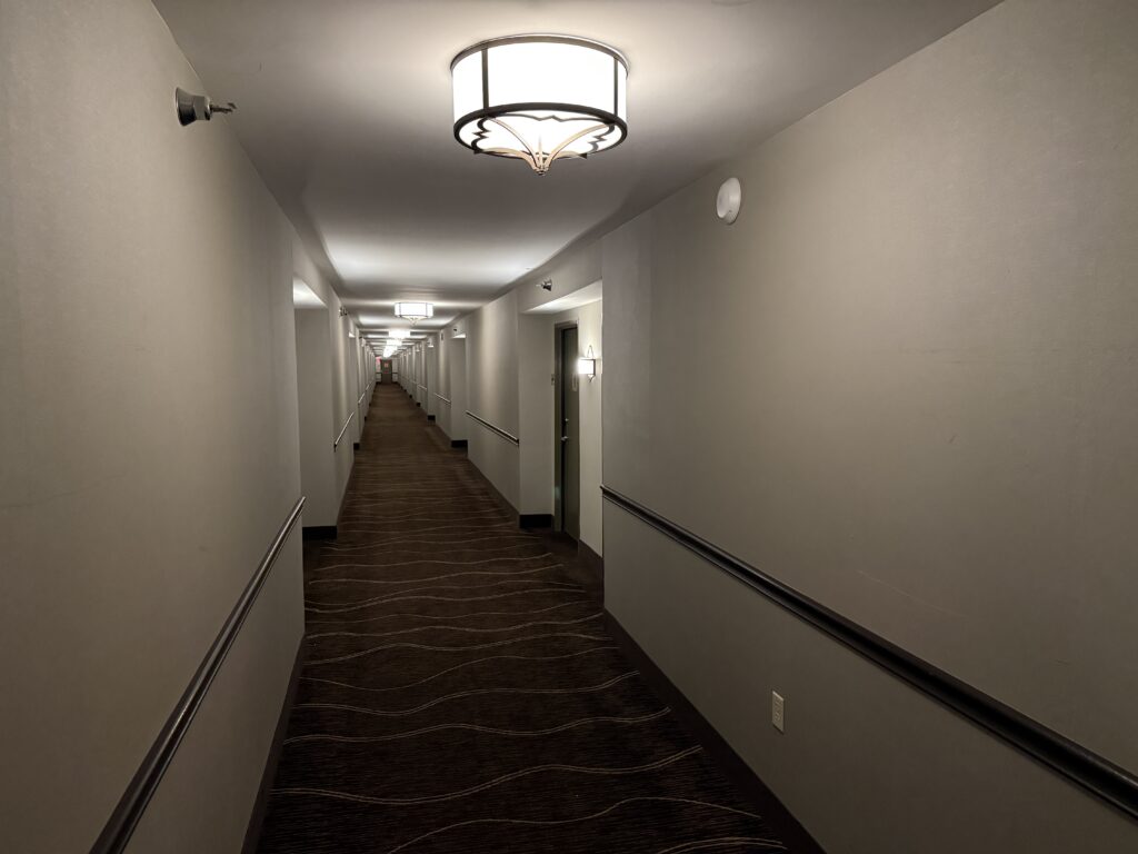 Long hotel hallway viewed from near foreground, beige walls, patterned carpet, and ceiling lights along the corridor.
