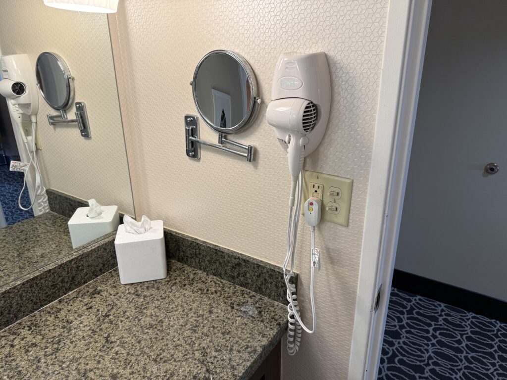 A makeup mirror on the wall next to a wall mounted hair dryer.