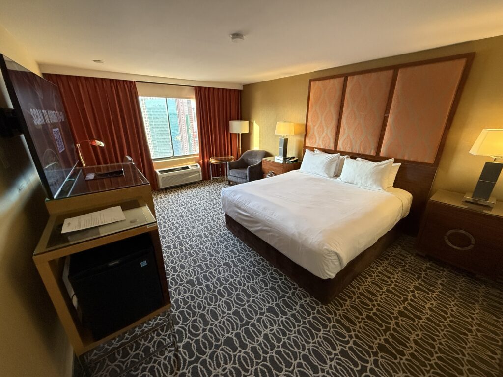 A wide angle view of the room with a king bed pushed against a padded headboard and a window in the background. 