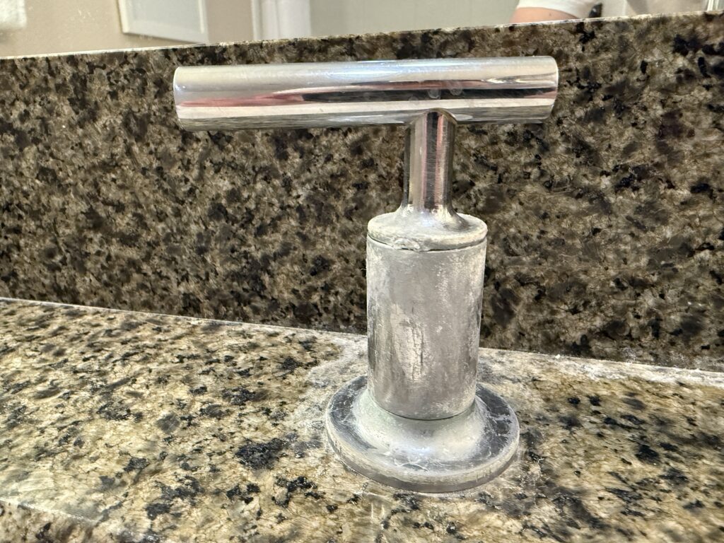 Close-up of a chrome faucet handle rencrusted with a whilte mineral buildup, mounted on a  granite countertop.