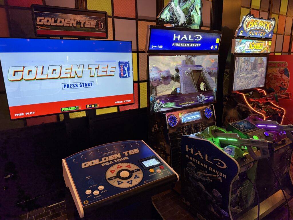 Row of arcade game machines with bright screens showing Golden Tee and Halo/Fireteam Raven titles in a dim, neon-lit arcade.