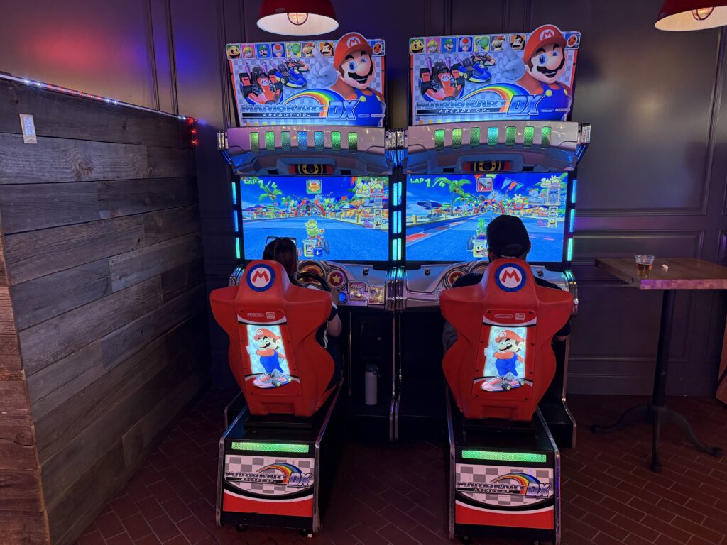 Two players in red Mario Kart DX arcade racing seats, facing bright screens with tropical track visuals.