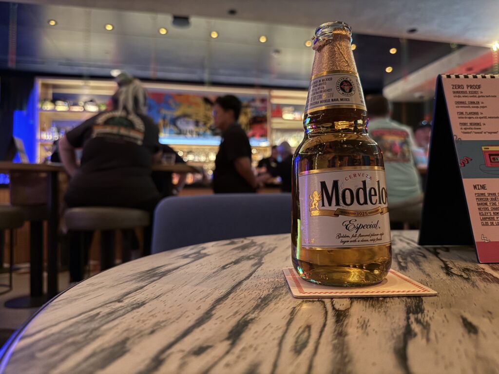 A bottle of Modelo sits on a table with an illuminated bar in the background. 