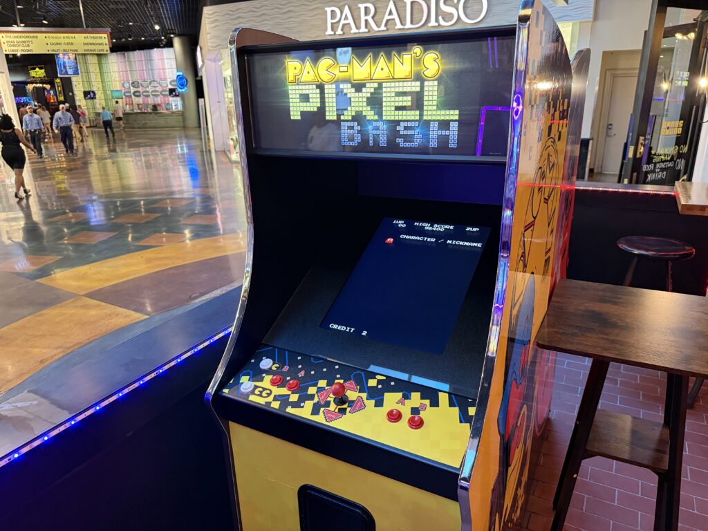 Pac-Man arcade cabinet in a shopping mall arcade, with game screen visible and people walking in the background