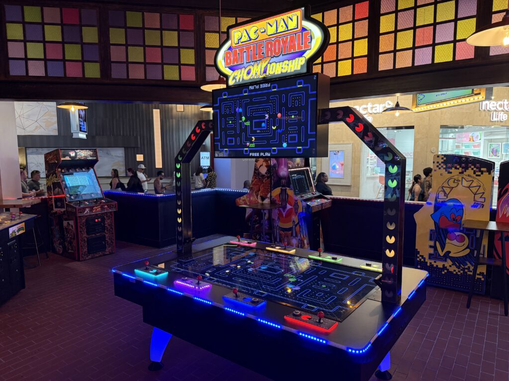 Pac-Man Battle Royale arcade setup with a large illuminated sign and a glowing blue maze screen in the foreground, players and other machines in the background.