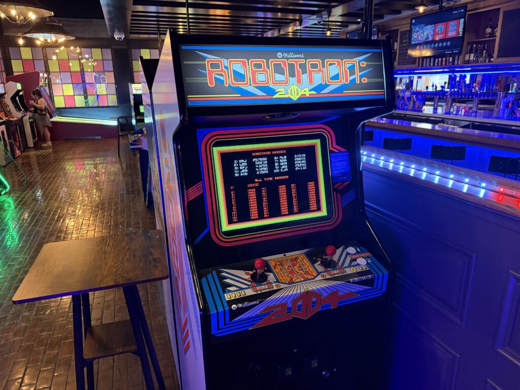 Retro arcade cabinet with bright neon graphics in a dimly lit bar, joystick controls and a large screen displaying bold 1980s-style typography.","Colorful arcade machine titled 'ABDOTRON 204' (or similar) with neon blue/pink accents, standing next to a wooden table in a neon-lit arcade bar.","Classic arcade game cabinet in a busy bar environment, featuring a large CRT screen and joystick controls amid blue LED lighting.","Slot of a retro arcade game on a tall machine in a bar, with vivid yellow, red, and blue artwork and a wooden table nearby.","Old-school arcade cabinet with neon artwork and a numeric screen, set in a lively bar with colorful wall panels and seating area."